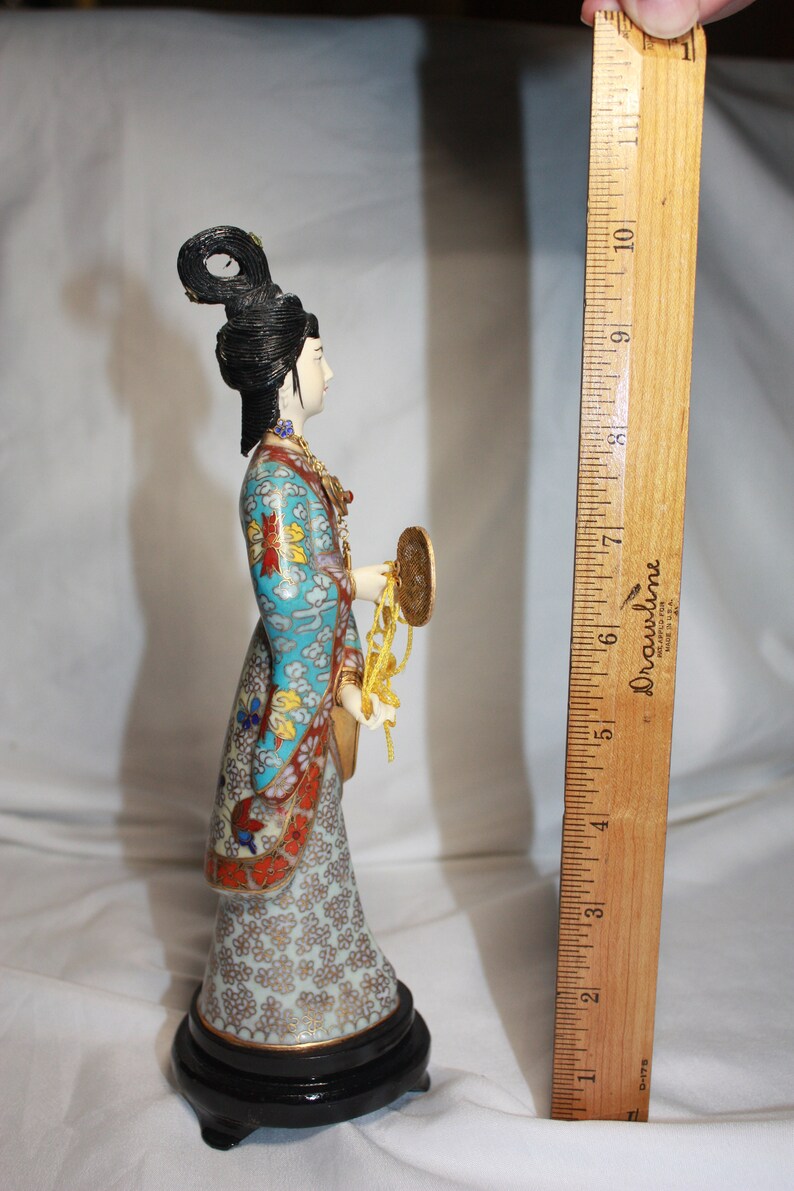 May include: A porcelain figurine of a woman in a blue and gold patterned robe, holding a golden fan and a string of beads. The figurine is standing on a black base.