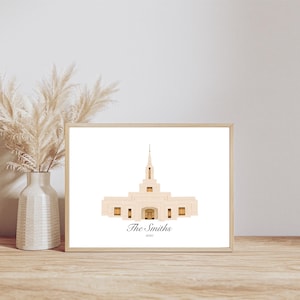 May include: A framed print featuring a beige building with a tall spire, labeled "The Smiths 2020". The artwork is displayed on a wooden surface, next to a white vase with dried ornamental grass.