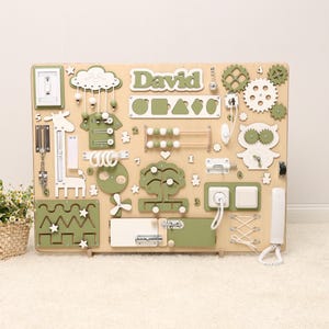 Può includere: Una tavola di attività in legno per bambini, con elementi interattivi come ingranaggi, interruttori e forme. La tavola è beige e verde oliva, con il nome "David". Include una giraffa e altri giocattoli educativi.