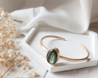 May include: A gold bangle bracelet with a green labradorite gemstone set in the centre. The bracelet is on a white surface.