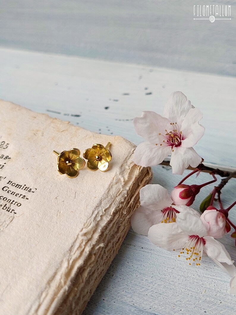 Op de afbeelding: Een paar gouden oorringen in de vorm van een bloem op een vintage boek met delicate roze bloesems.