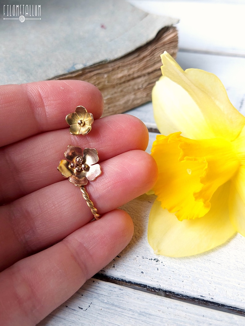 Op de afbeelding: Een paar gouden bloemoorbellen met een gedraaid draadontwerp. De oorbellen worden getoond op een hand tegen een witte houten achtergrond.