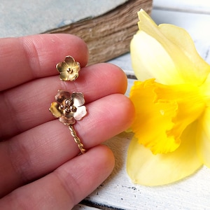 Op de afbeelding: Een paar gouden bloemoorbellen met een gedraaid draadontwerp. De oorbellen worden getoond op een hand tegen een witte houten achtergrond.