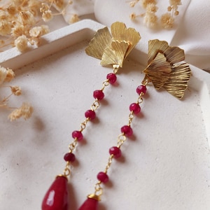 May include: Gold earrings with a layered leaf design and dangling red gemstone beads. The earrings are on a white surface.