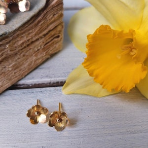 Op de afbeelding: Een paar gouden oorbelletjes in de vorm van een bloem. De oorbelletjes zijn klein en delicaat, met een simpel ontwerp. De oorbelletjes worden getoond tegen een witte houten achtergrond met een gele bloem.