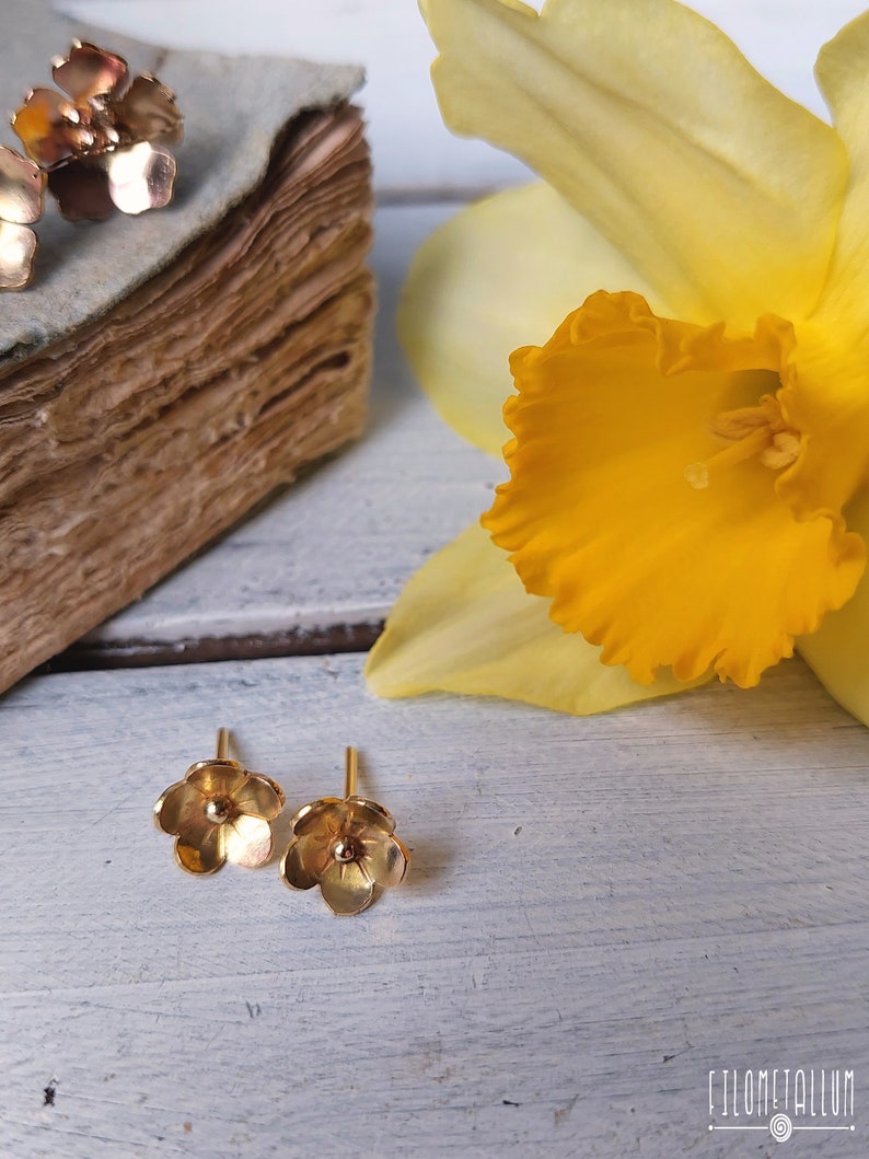 Op de afbeelding: Een paar gouden oorbelletjes in de vorm van een bloem. De oorbelletjes zijn klein en delicaat, met een simpel ontwerp. De oorbelletjes worden getoond tegen een witte houten achtergrond met een gele bloem.