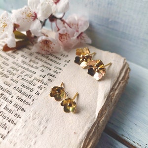 Op de afbeelding: Een paar gouden oorringen in de vorm van een bloem op een vintage boek. De oorringen zijn klein en delicaat, met een eenvoudig ontwerp.