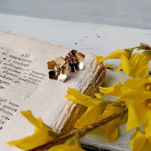 Op de afbeelding: Een paar gouden oorringen in de vorm van een bloem met kleine gouden kralen in het midden van elke bloem. De oorringen liggen op een boek met gele bloemen.