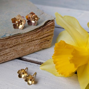 Op de afbeelding: Een paar gouden oorringen in de vorm van een bloem met een enkele parel in het midden van elke bloem. De oorringen worden getoond op een houten oppervlak met een gele bloem en een vintage boek.