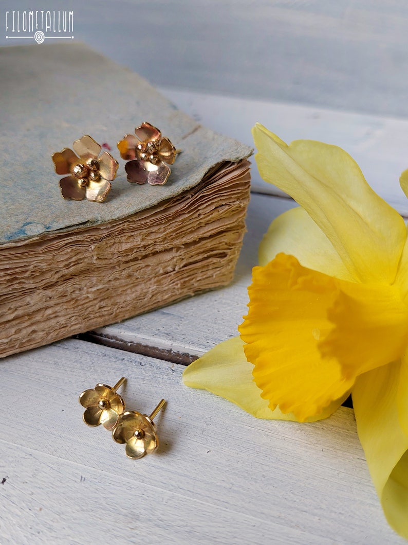 Op de afbeelding: Een paar gouden oorringen in de vorm van een bloem met een enkele parel in het midden van elke bloem. De oorringen worden getoond op een houten oppervlak met een gele bloem en een vintage boek.