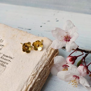 Op de afbeelding: Een paar gouden oorringen in de vorm van een bloem op een vintage boek met delicate roze bloesems.