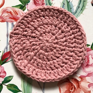 Op de afbeelding: Een ronde, roze gehaakte onderzetter. De onderzetter is gemaakt van zacht, getextureerd garen en heeft een delicate, handgemaakte uitstraling.