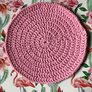 Può includere: Un sottopentola rotondo all'uncinetto rosa con una superficie testurizzata. Il sottopentola è realizzato con punto basso e ha un design circolare.