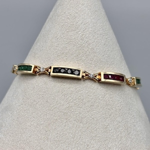 May include: A gold bracelet featuring rectangular links with alternating gemstones. The bracelet includes emerald green, black, and ruby red stones, along with small diamond accents. The bracelet is displayed on a neutral background.
