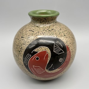 May include: A speckled ceramic vase with a round body and a green rim. The vase features a design of two fish, one red and one black, in a yin-yang style. The fish have white details.
