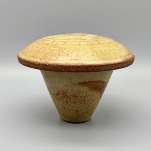 May include: A ceramic vessel with a wide, flat, mushroom-shaped top and a tapered, conical base. The piece features a speckled, light tan and cream glaze with a dark, textured brown rim around the edge of the top and earthy brown accents.