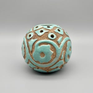 May include: A spherical ceramic vase with a textured, unglazed brown clay surface featuring raised, glossy light blue floral and vine patterns. The top of the vase has several circular openings integrated into the decorative design.