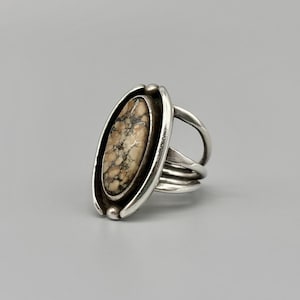 May include: A silver ring featuring an oval-shaped stone with a mottled beige and gray pattern. The stone is set in a silver bezel, and the ring has a split shank design with three bands.