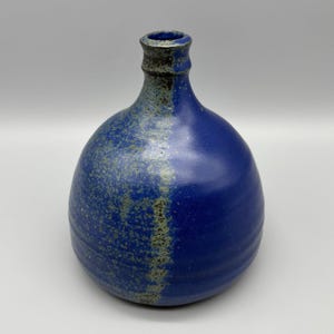 May include: A handmade ceramic vase with a rounded body and a narrow neck. The vase is primarily a deep blue color, with a textured, speckled, greenish-gray section on the left side and around the neck. The vase has a rustic, artisanal aesthetic.