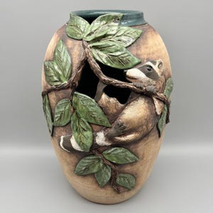 May include: A ceramic vase with a raccoon design. The vase is tan with a dark green rim and features a raccoon climbing through a tree with green leaves. The raccoon is brown, tan, and white. The leaves are a dark green color.