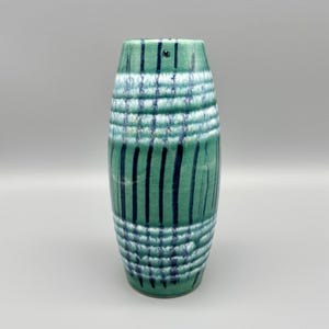 May include: A cylindrical ceramic vase with a green base color and a pattern of dark blue vertical stripes and horizontal lines. The vase has a textured surface and a slightly flared top. The vase is approximately 8 inches tall.
