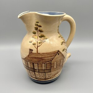 May include: A beige ceramic pitcher featuring a hand-painted brown illustration of a two-story wooden house with a chimney and a balcony. The pitcher has a curved handle, a wide spout, and a blue interior rim, with a dark blue band at the base.