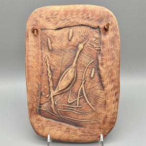 May include: A rectangular, brown ceramic plaque with a carved relief of a heron in a marsh setting. The heron is depicted with long legs and a curved neck, surrounded by reeds and water plants. The artist's signature is visible on the left side.