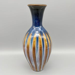 May include: A tall, decorative ceramic vase with a flared top and a rounded body. The vase features vertical stripes in shades of blue, brown, and gold. The top is dark blue and the rim is brown. The vase is a decorative home accent.