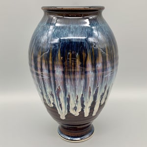 May include: A large, round ceramic vase with a dark brown base and a blue, white, and gold glaze. The glaze has a drip effect, creating a unique and artistic look.