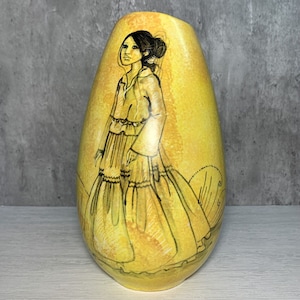 May include: A yellow ceramic vase with a hand-painted design of a woman in a long dress. The woman is depicted in black and white, and the vase has a yellow background with subtle orange and brown accents.