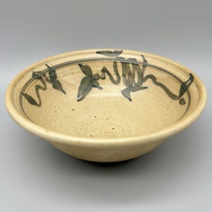 May include: A beige ceramic bowl with a dark green abstract design around the rim. The bowl has a wide, shallow shape and a speckled texture. The interior is a lighter beige color.