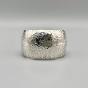 May include: A wide, silver-toned cuff bracelet featuring a distinct hammered texture across its entire outer surface. The metal has a reflective, polished finish that catches the light, creating varied highlights and shadows across the curved form.