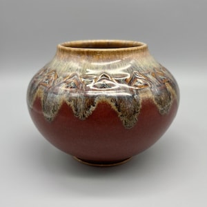 May include: A round ceramic vase with a wide opening. The vase has a reddish-brown base, transitioning to a lighter brown and beige rim. The upper portion features a textured, geometric pattern.