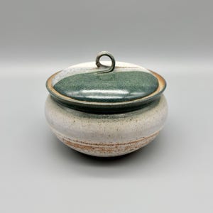 May include: A round ceramic lidded jar with a speckled off-white base and a band of earthy brown texture near the bottom. The lid features a glossy dark green glaze with a small, curved loop handle centered on top, creating a smooth finish.