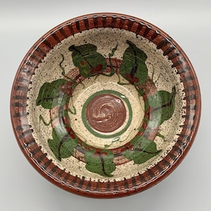 May include: A round ceramic bowl with a speckled cream-colored interior featuring a hand-painted pattern of green leaves and red berries. The center has a brown spiral design, and the wide, fluted rim is decorated with alternating brown stripes.
