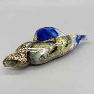 May include: A decorative glass sculpture in the shape of a seashell. The sculpture features a blend of colors, including gold, blue, green, and yellow. The glass has a glossy finish and is designed to resemble a natural seashell.