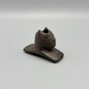 May include: A dark brown ceramic sculpture featuring a stylized cat-like head with pointed ears and a smooth, rounded face. The figure is attached to a flat, rectangular base with a textured, mottled surface finish and a small opening at the top.