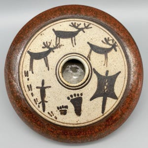 May include: Round ceramic vessel with a speckled off-white interior and a reddish-brown rim. The center features a circular opening. Black deer, footprints, and abstract shapes decorate the surface. The vessel has a rustic, handmade appearance.