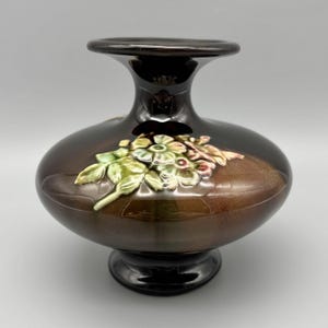 May include: A brown ceramic vase with a glossy finish. The vase features a bulbous body, a flared neck, and a pedestal base. A floral relief design with green, white, and pink accents adorns the front of the vase. The vase is approximately 8 inches tall.