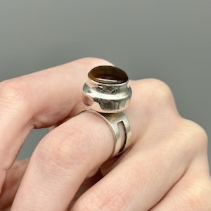May include: A silver ring with a large, oval, brown gemstone. The ring has a unique design with a wide band and a raised setting for the stone. The gemstone is a warm brown color, and the ring is set against a neutral background.