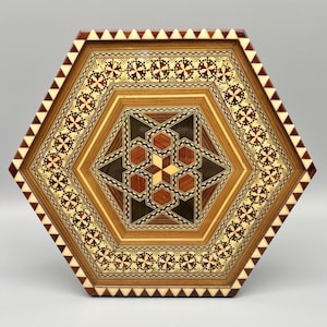 May include: A hexagonal wooden tray featuring intricate marquetry with geometric patterns. The center displays a star motif in shades of brown and cream, surrounded by a border of repeating floral-like shapes and a sawtooth edge in dark brown and white.