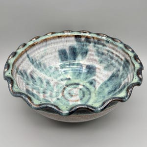 May include: A handmade ceramic bowl with a scalloped rim. The bowl features a blend of blue, green, and brown hues, with a spiral design in the center. The rim has a darker, textured finish.