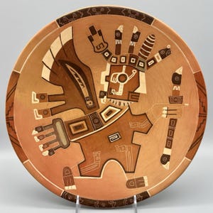 May include: A round, terracotta-colored ceramic plate featuring a stylized, geometric figure in shades of brown and cream. The central figure holds a staff and is surrounded by intricate patterns and designs. The plate's rim has a decorative border.