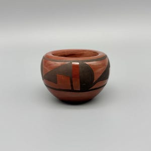 May include: A small, round, terracotta-colored pottery bowl with a dark brown geometric design. The bowl has a wide opening and a smooth, unglazed interior. The exterior features a repeating pattern of angular shapes.