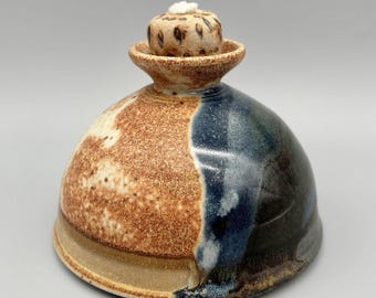 May include: A handcrafted ceramic oil lamp with a domed shape. The lamp is divided into two color sections: a speckled brown and a dark blue. A small, textured lid with a wick sits on top.