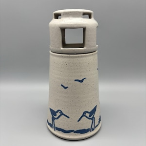 May include: A speckled ceramic lighthouse-shaped container with a square opening and a removable top. The container is decorated with blue bird and wave designs. The lighthouse has a speckled, off-white color.
