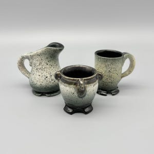 May include: Three speckled ceramic pieces: a small pitcher, a handled mug, and a small pot with handles. The pieces are in shades of gray and white with a speckled pattern. The pot has a dark brown rim.