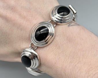 May include: Silver-toned bracelet featuring oval and circular links with black stone insets. The bracelet is composed of multiple connected segments, each with a polished, reflective surface. The design is symmetrical, with the black stones adding contrast to the silver metal.