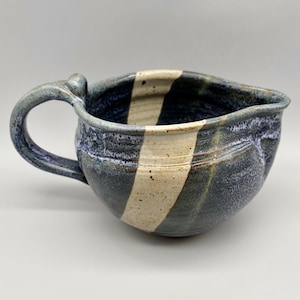 May include: Handmade ceramic pitcher with a handle and pouring spout. The pitcher features a dark blue and white striped design. The handle and rim have a speckled, textured appearance. The interior is dark blue.