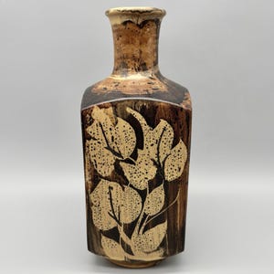 May include: A tall, brown ceramic vase with a rectangular body and a narrow neck. The vase features a leaf design in beige and black, with a textured surface. The vase is approximately 10 inches tall.